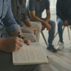 Man in group therapy writing in a notebook. Male therapist making notes on mental health behavioral progress and key talking points during a team building activity in an interactive support session